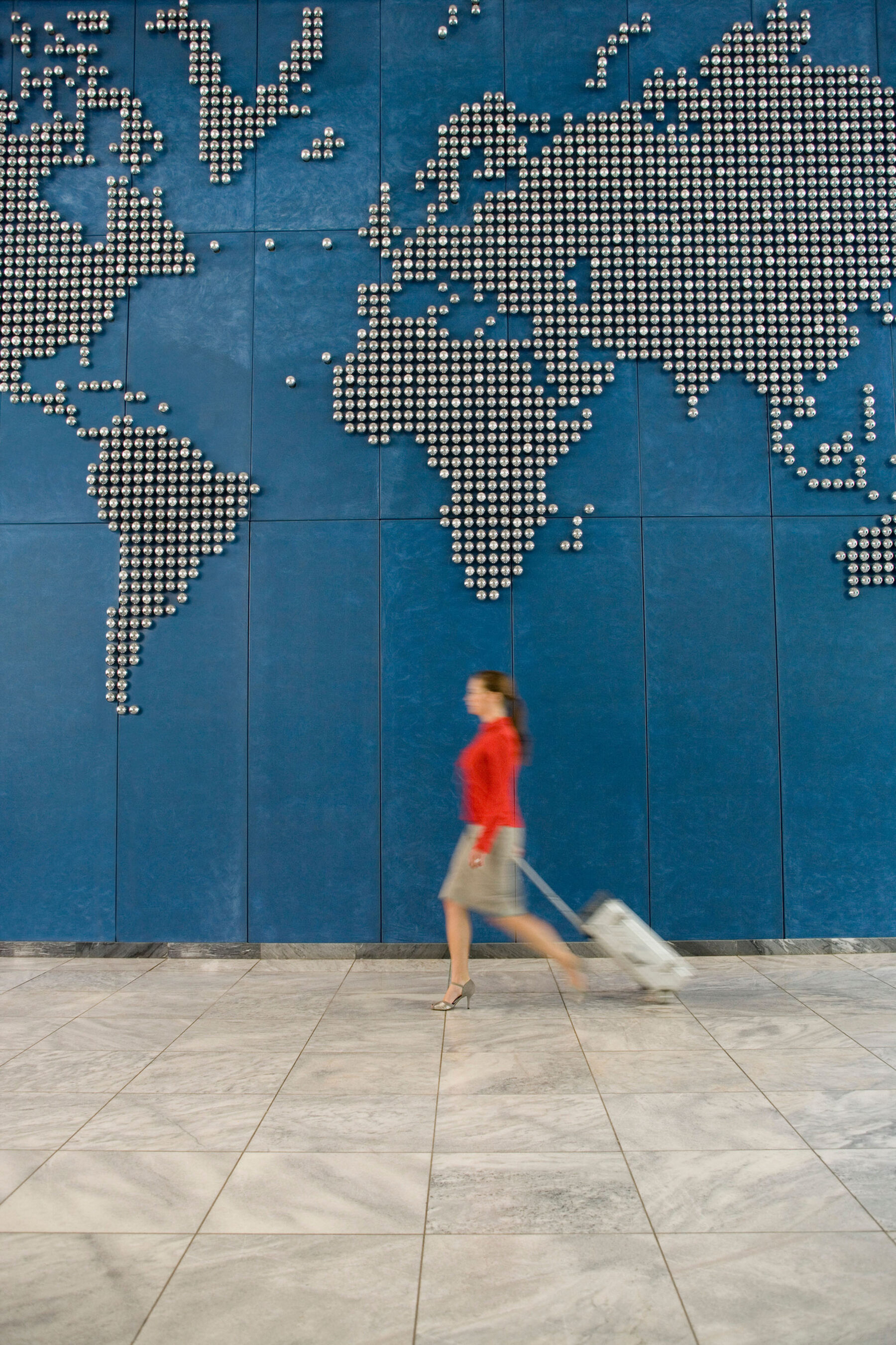 Woman walking past a wall with the world map on while pulling luggage behind her